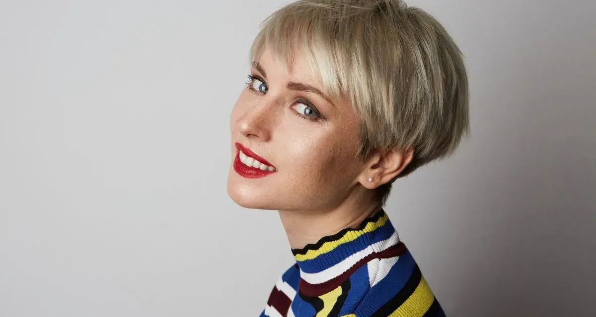 Kobieta z blond fryzurą pixie, która dodaje objętości cienkim włosom. Uśmiecha się, ma na sobie kolorowy sweter.