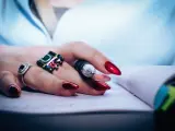 Dłoń z błyszczącymi, czerwonymi paznokciami i pierścionkami trzyma długopis nad notatnikiem. Dbałość o paznokcie to jak witaminy na paznokcie – efekt widać od razu.