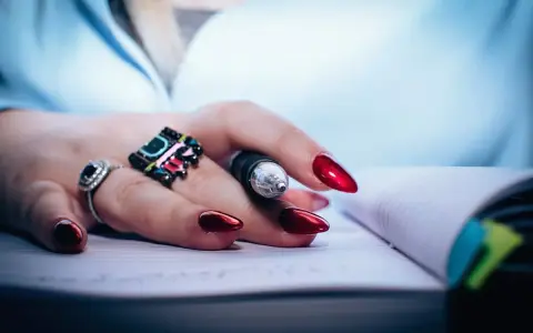 Dłoń z błyszczącymi, czerwonymi paznokciami i pierścionkami trzyma długopis nad notatnikiem. Dbałość o paznokcie to jak witaminy na paznokcie – efekt widać od razu.