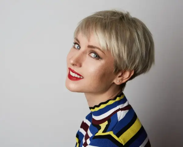 Kobieta z blond fryzurą pixie, która dodaje objętości cienkim włosom. Uśmiecha się, ma na sobie kolorowy sweter.