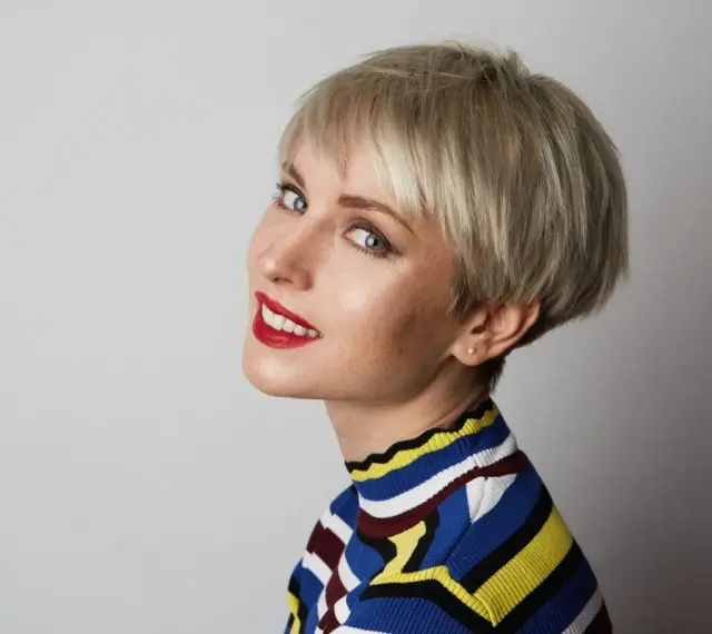 Kobieta z blond fryzurą pixie, która dodaje objętości cienkim włosom. Uśmiecha się, ma na sobie kolorowy sweter.
