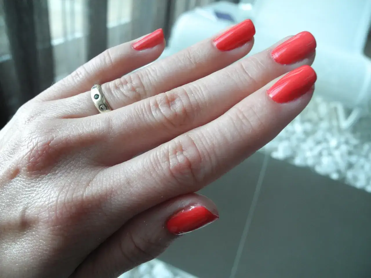 Dłoń z paznokciami pomalowanymi na jaskrawy koralowy kolor. Na serdecznym palcu widoczny pierścionek. To efekt pięknego manicure biologicznego.